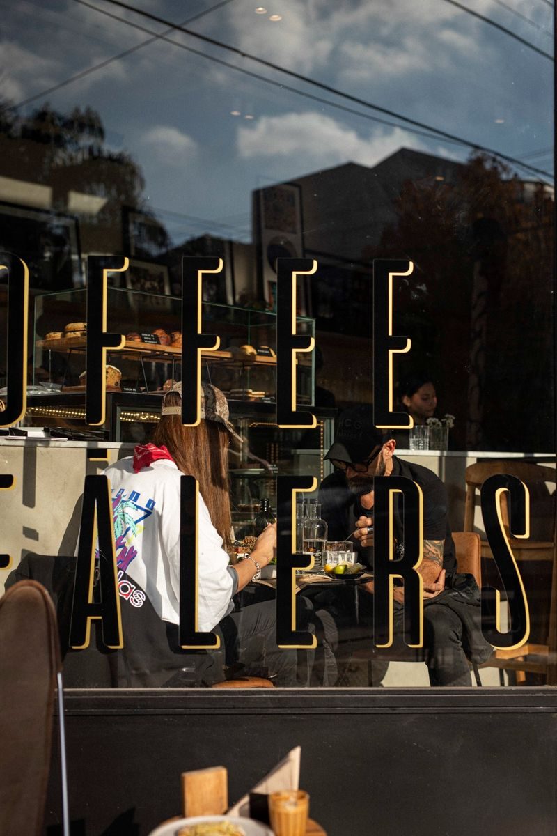 Vidriera Coffee Dealers SĀNTAL Colegiales — letras doradas en vidrio con clientes adentro y vitrina de pasteles