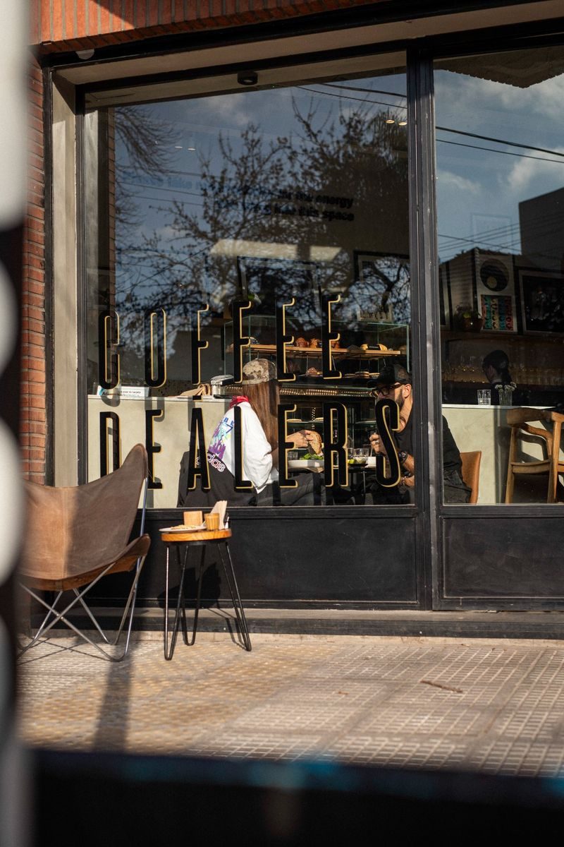 Fachada SĀNTAL Colegiales desde afuera — silla mariposa de cuero, vidriera Coffee Dealers y clientes en brunch