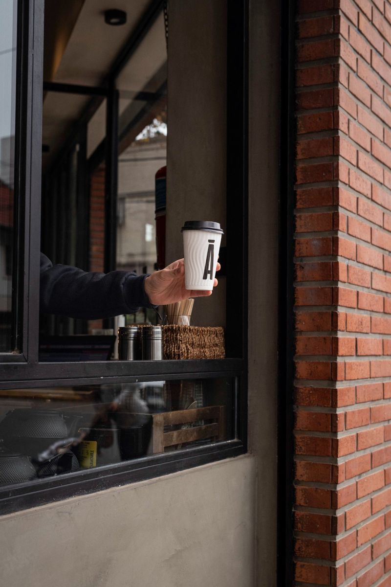 Mano entregando vaso takeaway SĀNTAL con logo Ā — ventana de ladrillos y entrega de café to go en Colegiales
